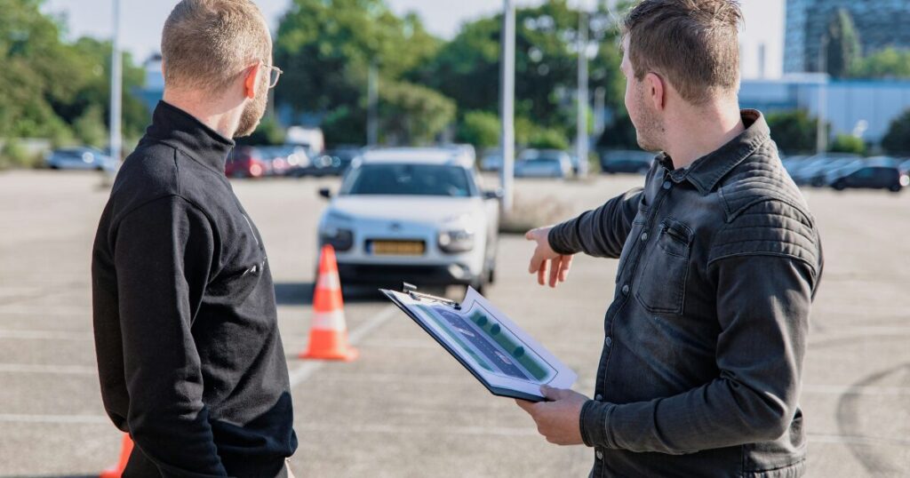 教習コースで、クリップボードを持った指導員が車を指さしながら説明している様子